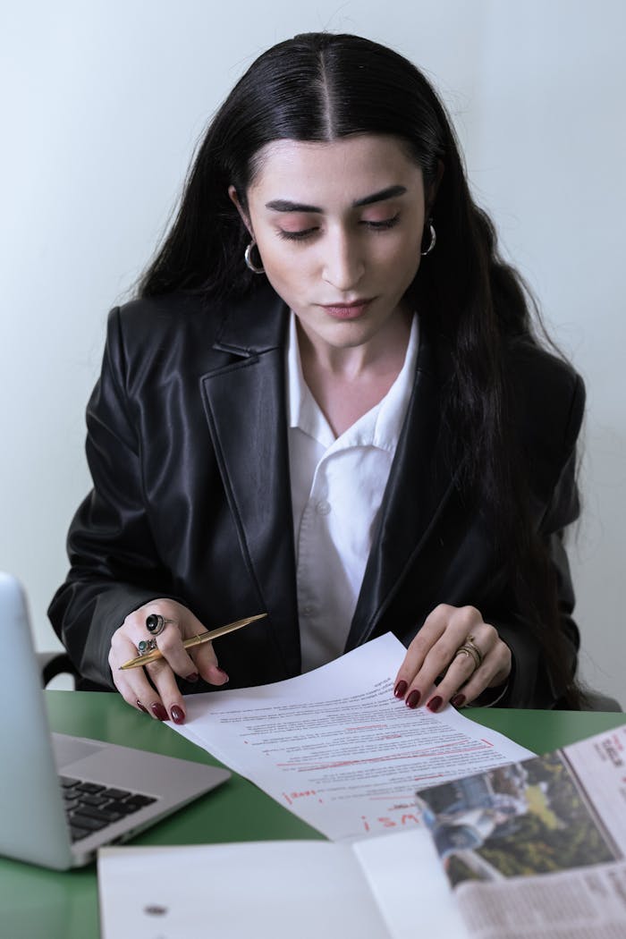 Professional woman in a leather coat reviewing documents next to a laptop in an office.